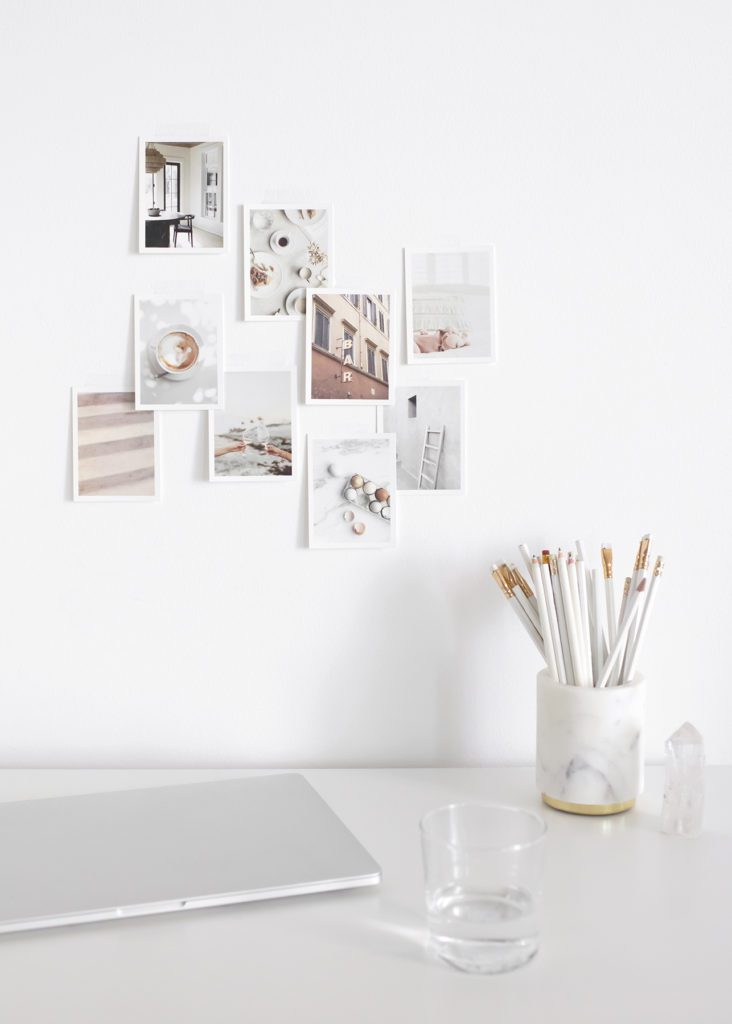 Image is white in tones, and has 9 polaroid's of various pictures hanging on a white wall. There is a desk with a closed silver laptop and a marble white pencil holder with a dozen white pencils.