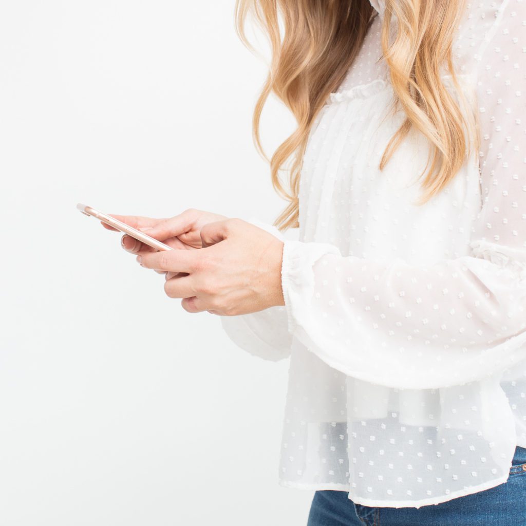 Photo of a woman in a white lace shirt, she is holding a phone. This photo show calling someone who cares as self-care. 