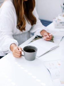 Image of woman writing in journal with coffee beside her - this shows chronic illness symptom tracker.