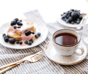 A picture of a tea cup with red tea representing red raspberry tea as one of the Best tea for menstrual cramps. It's on a plaid table cloth with blueberry pastry on a plate beside. 