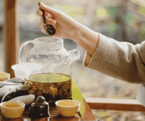 Image of a glass tea kettle with tea leaves in it. There are small tea cups beside the tea kettle and some loose tea leaves off the the side of the image. 