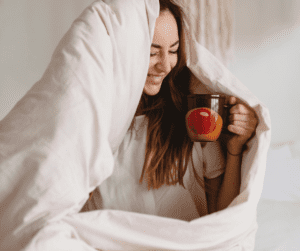 Image of a woman with a tea cup who has a fluffly duvet wrapped aroung her. She's sitting in bed with tea showing the best tea for menstrual pain to help with period pain. 