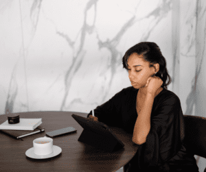 A woman sitting at a round table with an tablet and a stylus, shes working with notebooks and coffee as well on the table. This image is showing a woman working to grow your email list.