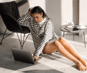 Image of a woman sitting on the floor typing on a laptop that is also on the floor. She is leaning her arm on a chair for support.