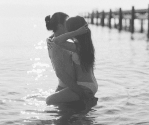 Image of a couple in water near a pier, the photo is in black and white and the man is holding the woman who has her legs wrapped around his waist.