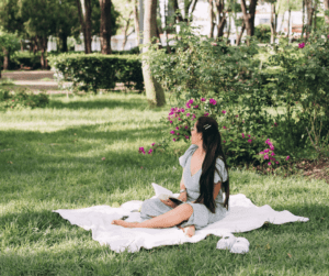 This image is a lady sitting on a blanket in the grass enjoying the beautiful summer weather. She has a notebook open on her lap to take down some unique blog ideas for summer content. 