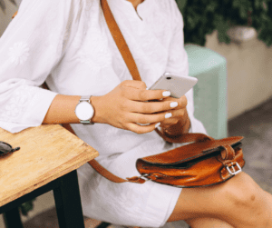 Image of a woman sitting on a wooden bench in a white sundress. She has a brown leather purse in her lap and a phone in her hand noting down unique blog ideas for summer. 