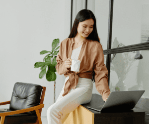 Image of a woman sitting on the edge of a table with a coffee mug in one hand. She is typing on a laptop with her other hand. 