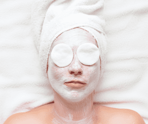 Image of a woman laying with cotton pads over her eyes with a white facial applied to the face. 